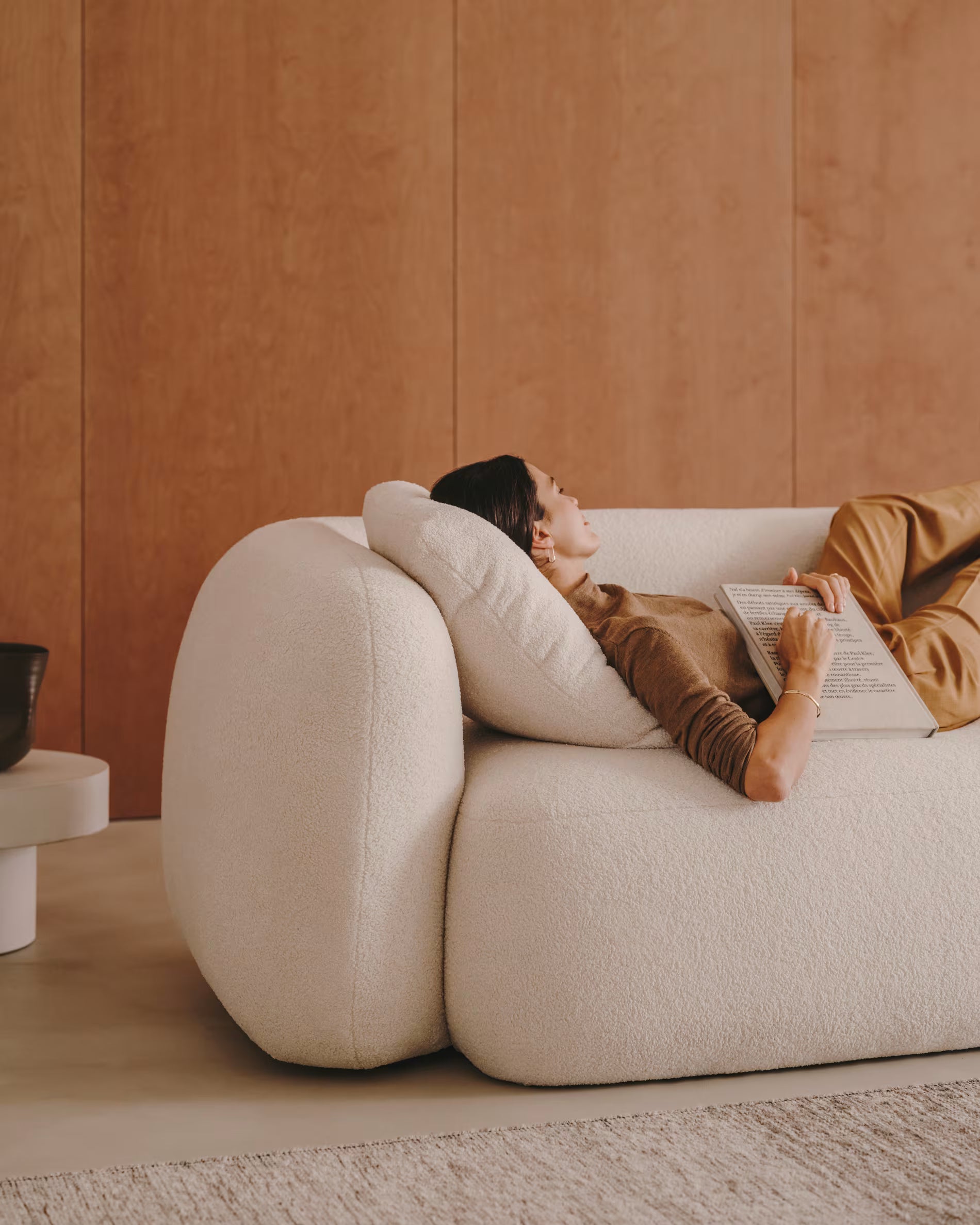 Person lying on a modern beige sofa reading a book against a wooden wall.