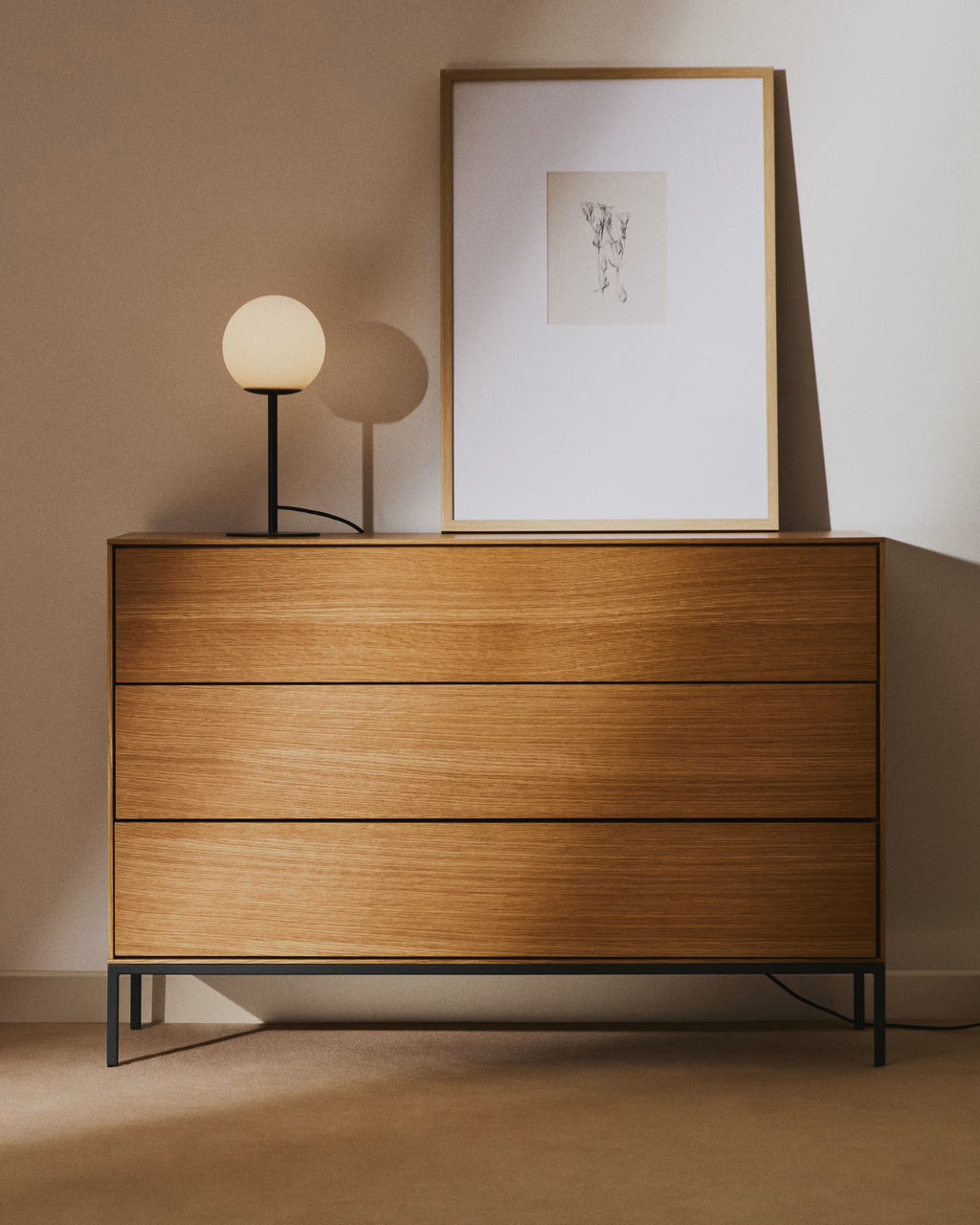 Wooden dresser with a framed picture and small lamp against a beige wall.